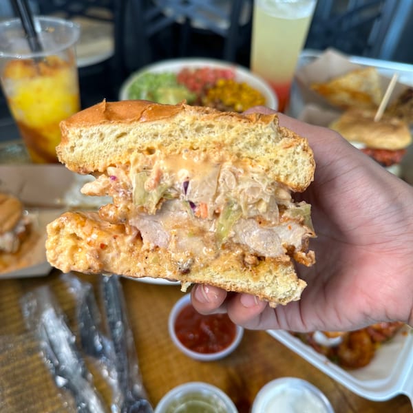 Hand holding a halved Korean fried chicken sandwich with slaw and sauce, with drinks and food in background.
