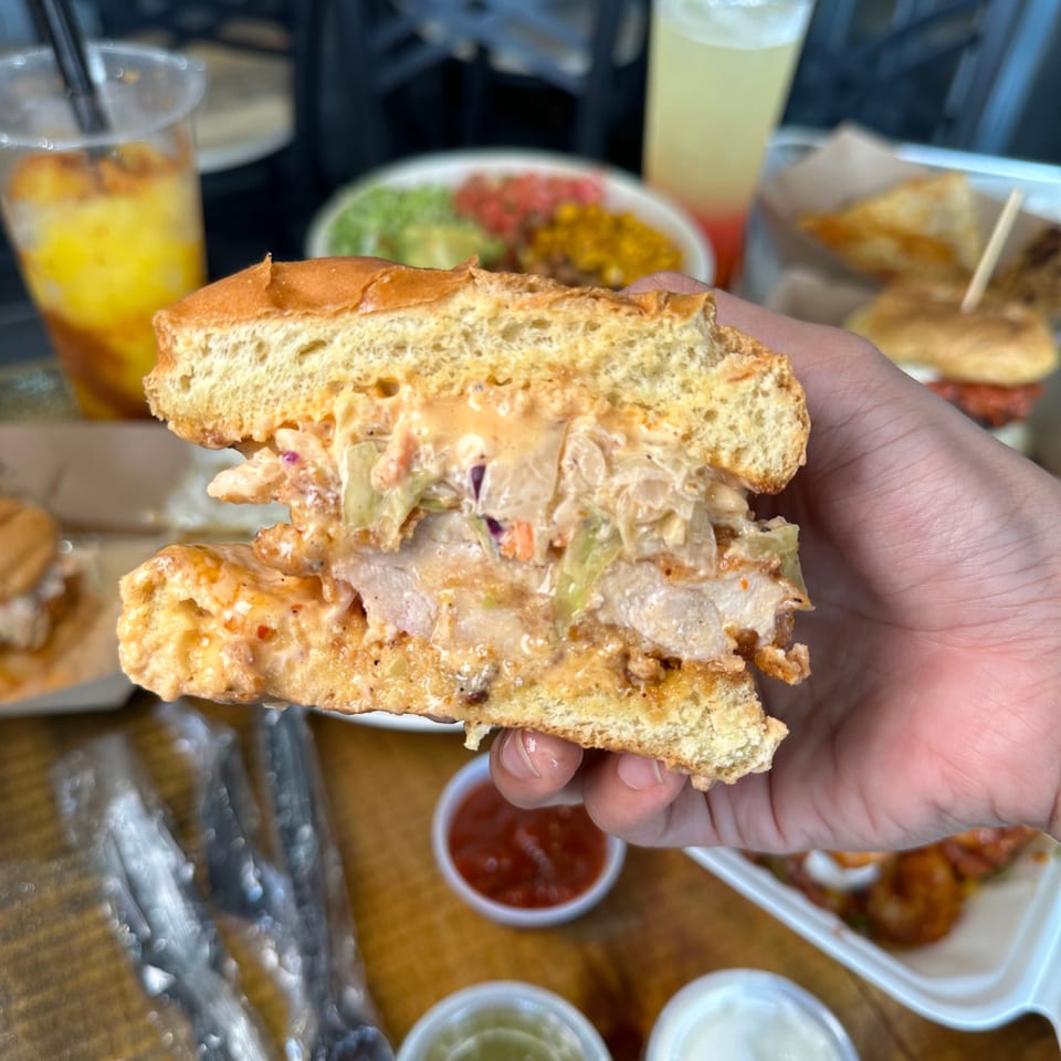 Hand holding a halved Korean fried chicken sandwich with slaw and sauce, with drinks and food in background.