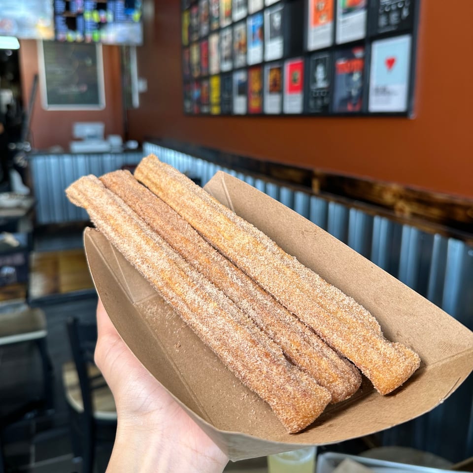 A hand holds a paper tray of four cinnamon sugar churros inside Bebop Korean Mexican Grill.