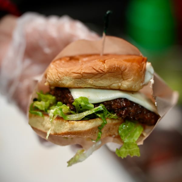A gloved hand holds a Korean burger with melted cheese, lettuce, and a beef patty.