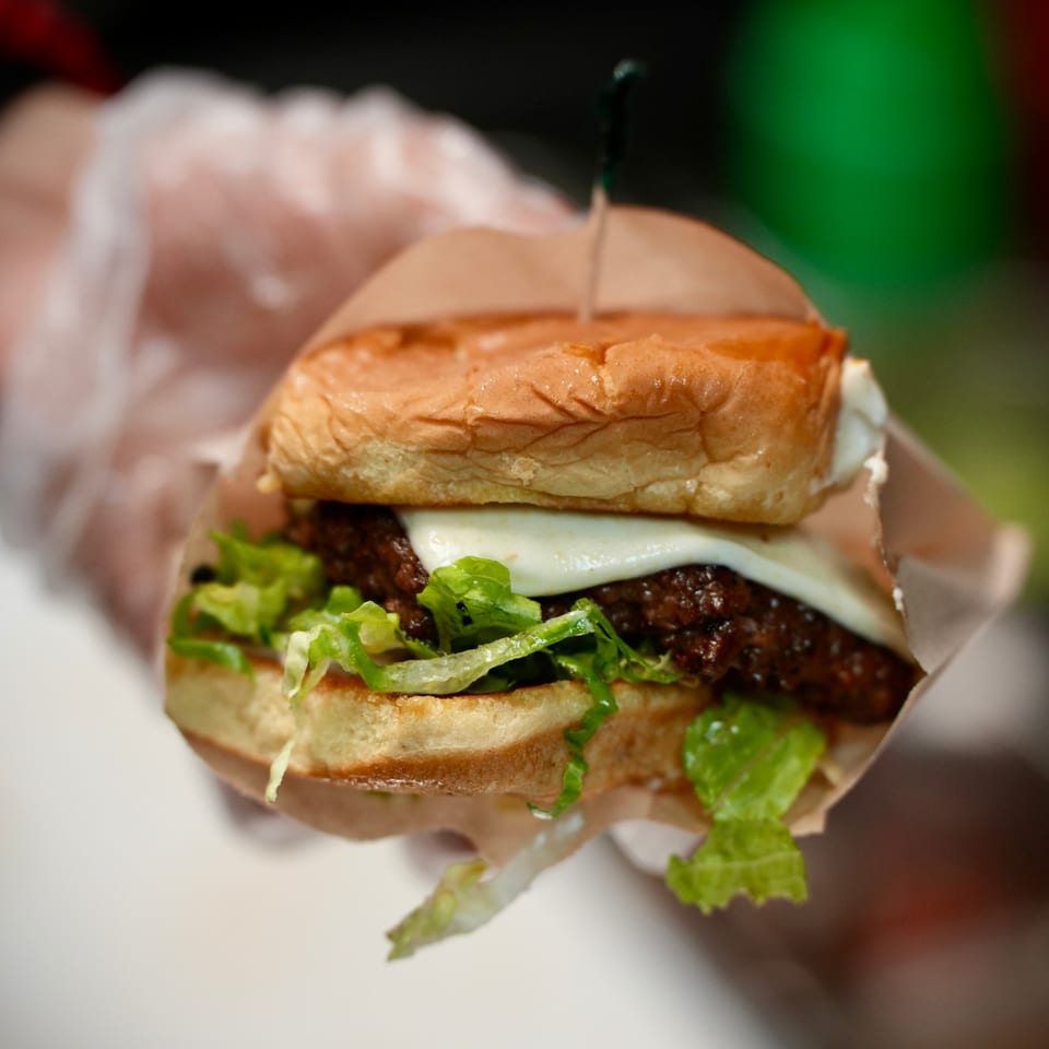 A gloved hand holds a Korean burger with melted cheese, lettuce, and a beef patty.
